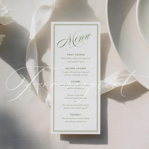 May include: A rectangular menu card with a green border and elegant script text. The menu lists courses such as butter lettuce salad, creamy mushroom soup, basil chicken, and pan-seared salmon. The card is on a white surface with a white ribbon.