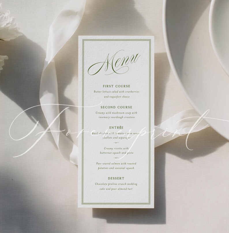 May include: A rectangular menu card with a green border and elegant script text. The menu lists courses such as butter lettuce salad, creamy mushroom soup, basil chicken, and pan-seared salmon. The card is on a white surface with a white ribbon.