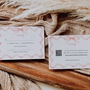May include: Two white RSVP cards with pink ribbon designs. The card on the left has a checkbox for "Joyfully accepts" and "Regretfully declines". The card on the right has a QR code and the text "to RSVP and for more details scan the QR code or visit our wedding website: www.audreyandiames.theknot.com". Both cards have the text "kindly respond by June st".