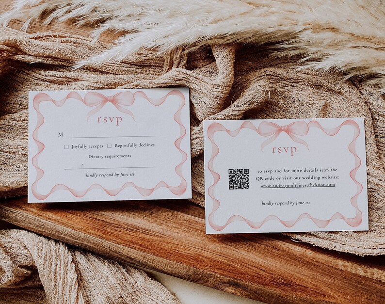 May include: Two white RSVP cards with pink ribbon designs. The card on the left has a checkbox for "Joyfully accepts" and "Regretfully declines". The card on the right has a QR code and the text "to RSVP and for more details scan the QR code or visit our wedding website: www.audreyandiames.theknot.com". Both cards have the text "kindly respond by June st".