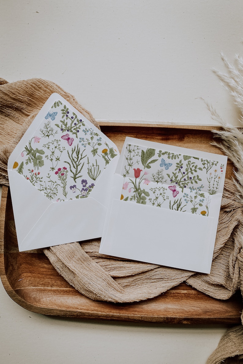 May include: Two white envelopes with a floral pattern on the front. The envelopes are on a wooden tray with a brown fabric underneath.