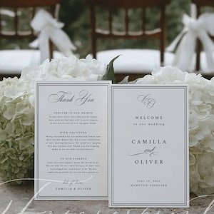 K&ouml;nnte beinhalten: Zwei wei&szlig;e Hochzeitskarten mit schwarzem Text. Auf einer steht "Danke" und auf der anderen "Willkommen zu unserer Hochzeit" mit den Namen Camilla und Oliver. Die Karten sind von wei&szlig;en Blumen umgeben.