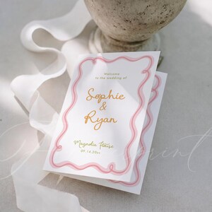 May include: A white wedding invitation card featuring a pink wavy border design. The text reads: Welcome to the wedding of Sophie & Ryan, Magnolia House, 09.14.20xx. The names Sophie and Ryan are printed in a gold script font on the centre of the card.