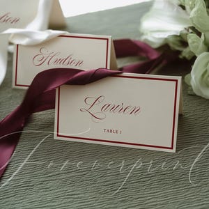 May include: Two folded cream-colored place cards with a dark red border and dark red cursive text. One card reads "Lauren" and "TABLE 1", while the other reads "Hudson". A deep burgundy ribbon is draped across the cards, and white flowers are visible.