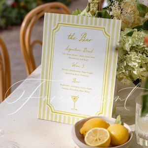 May include: A rectangular bar menu sign with yellow and white vertical stripes, featuring the text "the Bar," "Signature Drink," "Wine," and "Beer" with a martini glass illustration, placed next to a small bowl containing three yellow lemons.