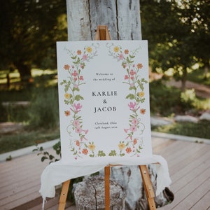 May include: A white wedding sign with a floral border. The sign reads "Welcome to the wedding of Karlie & Jacob, Cleveland, Ohio, 14th August 20XX".