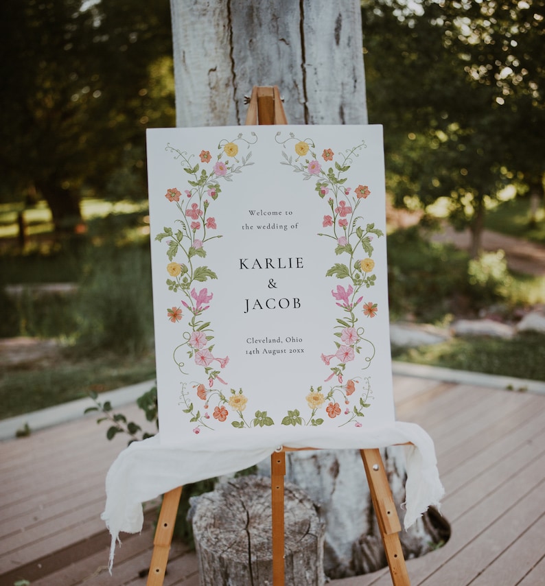 May include: A white wedding sign with a floral border. The sign reads "Welcome to the wedding of Karlie & Jacob, Cleveland, Ohio, 14th August 20XX".