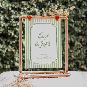 May include: A copper-colored metal sign holder with a green and white striped sign that reads "Cards & Gifts". The sign has a decorative border and is adorned with dried flowers and red velvet accents. The sign is for a wedding or event.