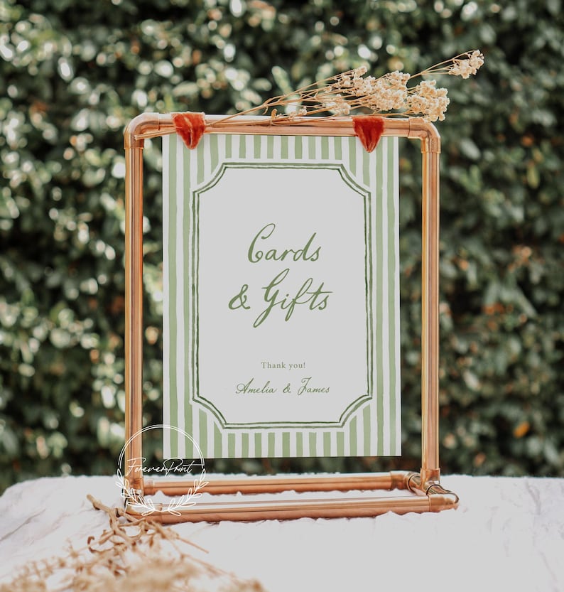 May include: A copper-colored metal sign holder with a green and white striped sign that reads "Cards & Gifts". The sign has a decorative border and is adorned with dried flowers and red velvet accents. The sign is for a wedding or event.