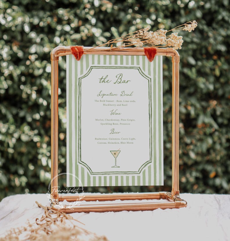 May include: A copper-colored metal bar sign with a green and white striped background. The sign displays a menu with categories for signature drinks, wine, and beer. Dried flowers and red velvet accents adorn the top.