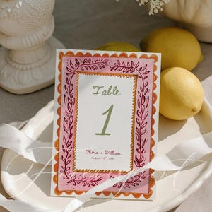 Puede incluir: Una tarjeta con el número de mesa con un borde rosa y naranja, que muestra la palabra "Table" y el número "1" en verde. La tarjeta incluye los nombres "Olivia + William" y la fecha "14 de agosto de 20xx".