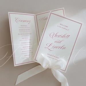 May include: A folded wedding programme with a white cardstock base and dusty rose border. The front features the text Welcome to the wedding of Scarlett and Lincoln, Palm Springs, August 25th, 20XX. A white silk ribbon is tied at the bottom.