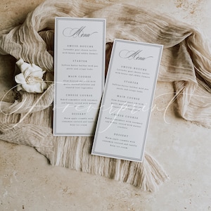 May include: Two rectangular menu cards with a dark blue border and elegant script. The cards are on a beige fabric with a white flower. The menu includes courses like Amuse-bouche, Starter, Main Course, Cheese Course, and Dessert.