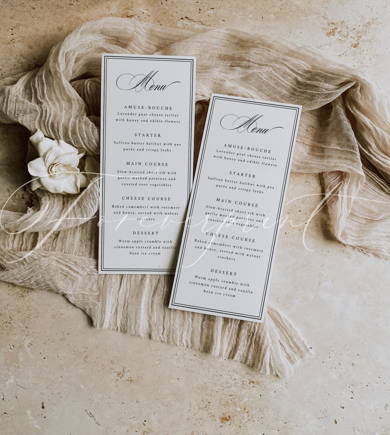 May include: Two rectangular menu cards with a dark blue border and elegant script. The cards are on a beige fabric with a white flower. The menu includes courses like Amuse-bouche, Starter, Main Course, Cheese Course, and Dessert.