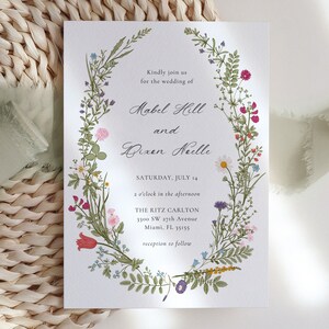 May include: Wedding invitation featuring a floral wreath design. The invitation includes the names Mabel Hill and Dixen Neille, along with the date, time, and location: The Ritz Carlton, Miami, FL. The wreath is composed of various colorful flowers and greenery.