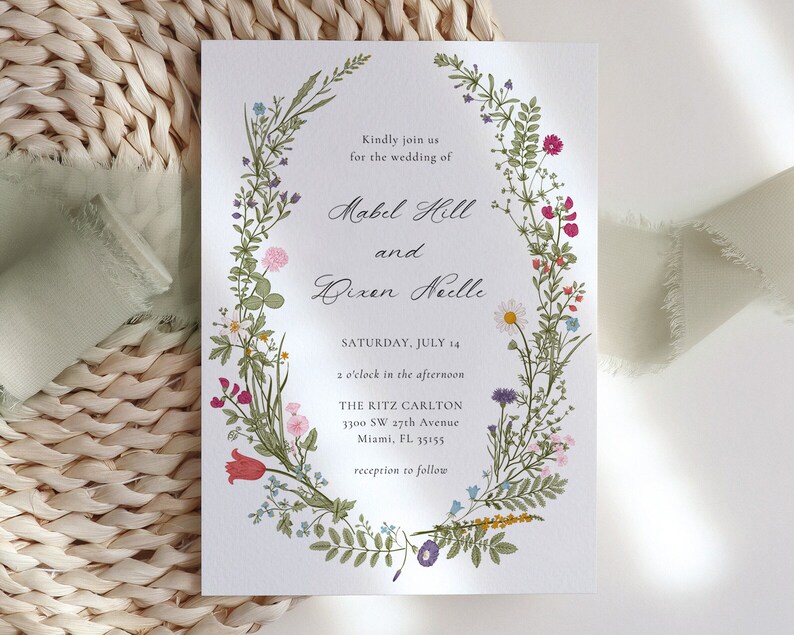 May include: Wedding invitation featuring a floral wreath design. The invitation includes the names Mabel Hill and Dixen Neille, along with the date, time, and location: The Ritz Carlton, Miami, FL. The wreath is composed of various colorful flowers and greenery.