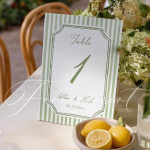 May include: A table number card with a green and white striped border. The card reads "Table 1" in green script, with the names "Chloe & Nick" and the date "08.13.20xx" below. A small bowl of lemons sits in front of the card.