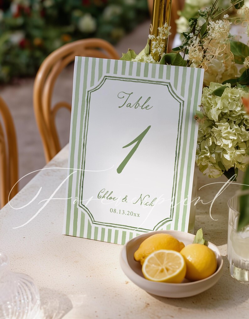 May include: A table number card with a green and white striped border. The card reads "Table 1" in green script, with the names "Chloe & Nick" and the date "08.13.20xx" below. A small bowl of lemons sits in front of the card.