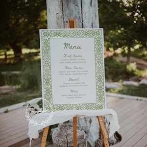 Può includere: Un tableau di menu bianco con un bordo verde e un menu scritto a mano. Il menu include un antipasto, un piatto principale, un dessert e opzioni di vino. Il menu è visualizzato su un cavalletto di legno.