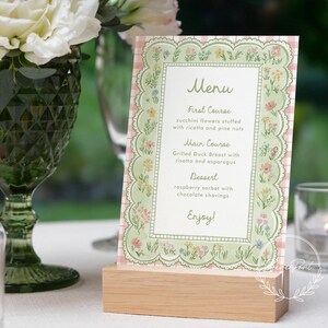 May include: A floral menu card with a scalloped edge, displayed on a wooden block. The menu features a pink and green floral design with the word "Menu" at the top. The menu lists the courses: First Course, Main Course, and Dessert.