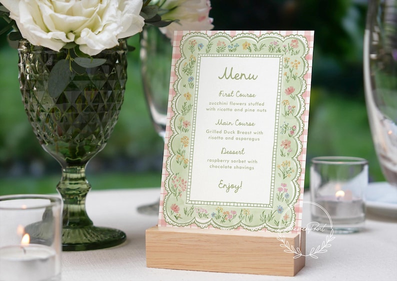 May include: A floral menu card with a scalloped edge, displayed on a wooden block. The menu features a pink and green floral design with the word "Menu" at the top. The menu lists the courses: First Course, Main Course, and Dessert.