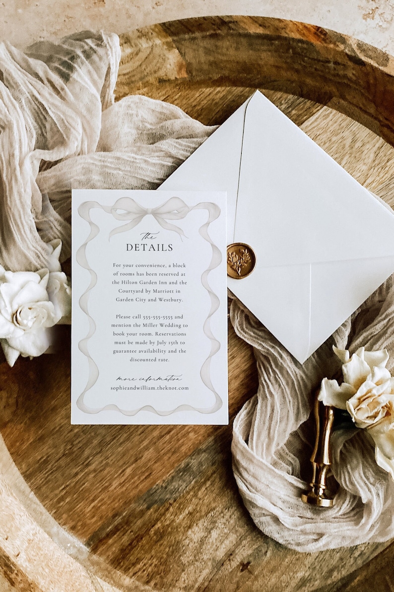 May include: A white card with a decorative border and the text "The Details" in a script font. The card provides information about a wedding, including the hotel block reservation details and contact information. The card is on a wooden surface with a white envelope and a gold wax seal.