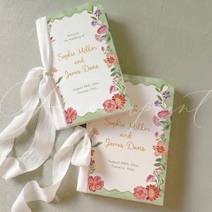 May include: Two rectangular wedding programs with white centers and sage green scalloped borders, decorated with watercolor flowers. The text reads: Welcome the Wedding of Sophie Miller and James Davis, August 28th, 20xx, Tuscany, Italy. Each has a white ribbon.
