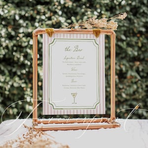 May include: A copper-colored metal framed sign with a pink and white striped background. The sign reads "the Bar" and lists signature drinks, wine, and beer options. Dried flowers and green velvet accents adorn the top of the frame, suitable for a wedding or event.