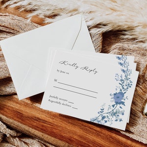 May include: A white RSVP card with blue floral accents. The card has the text "Kindly Reply by June 1st" and lines for guests to indicate if they will joyfully accept or regretfully decline the invitation.
