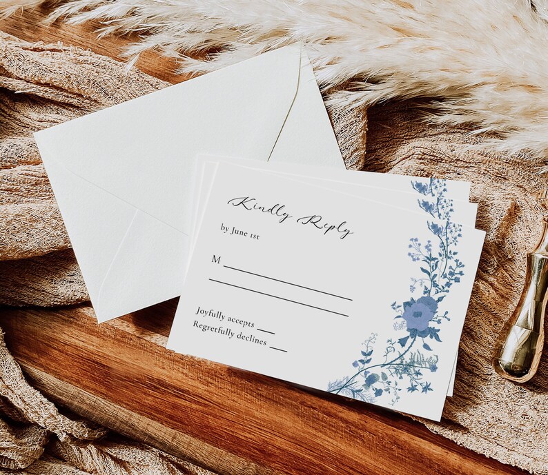May include: A white RSVP card with blue floral accents. The card has the text "Kindly Reply by June 1st" and lines for guests to indicate if they will joyfully accept or regretfully decline the invitation.