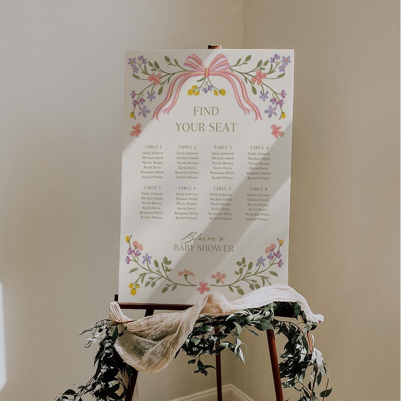 May include: A white seating chart with a pink bow and floral accents. The chart lists tables and names for a baby shower. The text reads "Find Your Seat" and "Blein's Baby Shower".