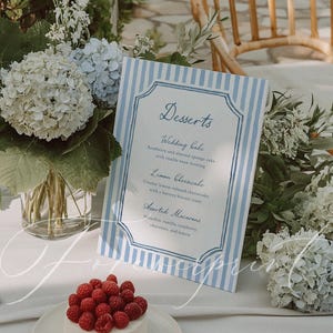 Könnte beinhalten: Eine Dessert-Tischdekoration mit einem kleinen weißen Kuchen mit frischen Himbeeren, einer Speisekarte mit blauen Streifen und Blumengestecken. Die Speisekarte listet Hochzeitstorte, Zitronenkäsekuchen und verschiedene Macarons auf.