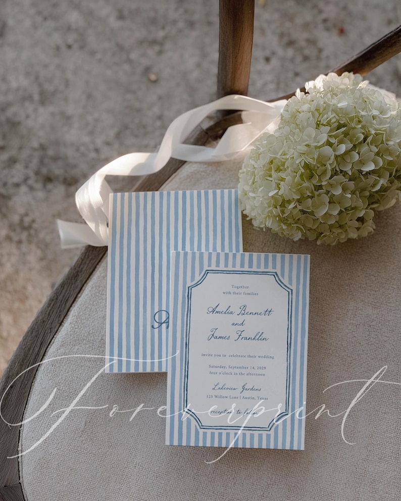 May include: Wedding invitation suite with blue and white striped design. The invitation includes the names Amelia Bennett and James Franklin, along with event details. A white ribbon and a bouquet of white flowers are also present.