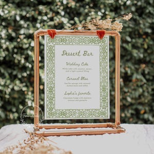 May include: A copper pipe stand with a white card featuring a green patterned border and the text "Dessert Bar". The card lists the wedding cake, caramel bliss, and Sophie's favorite desserts.