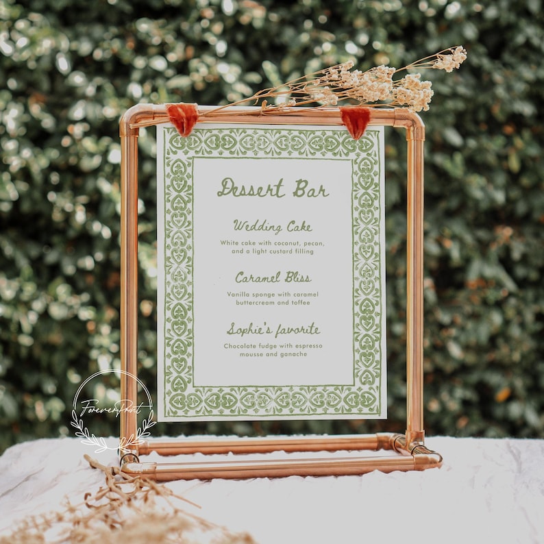 May include: A copper pipe stand with a white card featuring a green patterned border and the text "Dessert Bar". The card lists the wedding cake, caramel bliss, and Sophie's favorite desserts.