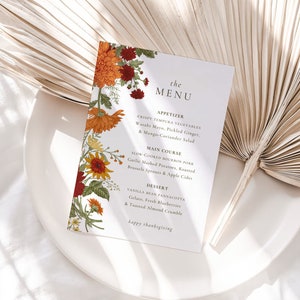 Pu&ograve; includere: Un menu del Ringraziamento con un design floreale con fiori arancioni, rossi e gialli. Il menu include opzioni di antipasti, piatti principali e dessert. Il testo recita "the MENU", "APPETIZER", "CRISPY TEMPURA VEGETABLES", "Wasabi Mayo, Pickled Ginger, & Mango-Coriander Salad", "MAIN COURSE", "SLOW COOKED BOURBON PORK", "Garlic Mashed Potatoes, Roasted Brussels Sprouts & Apple Cider", "DESSERT", "VANILLA BEAN PANNACOTTA", "Gelato, Fresh Blueberries & Toasted Almond Crumble", "happy thanksgiving".