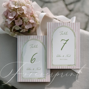 May include: Two wedding table number cards with pink and white stripes. Each card has a gold-framed arch with the word "Table" and a number in green. The names "Chloe & Nick" and the date are below.