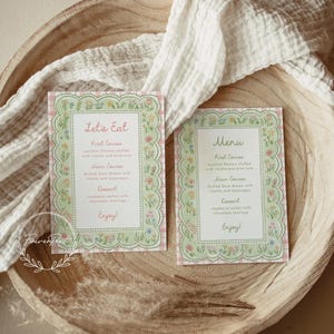May include: Two floral-bordered menu cards with the text "Let's Eat" and "Menu" on a wooden bowl. The cards have a pink and green color scheme with a floral design. The menu includes first course, main course, and dessert options.