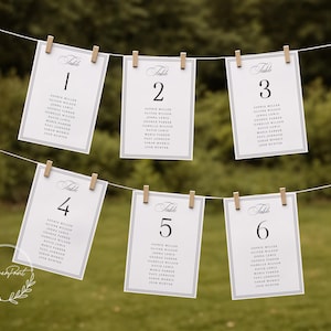 May include: Six white table number cards, numbered 1 through 6, are suspended from a string with wooden clothespins. Each card features a black border, elegant script, and a list of names. The background is a blurred green.