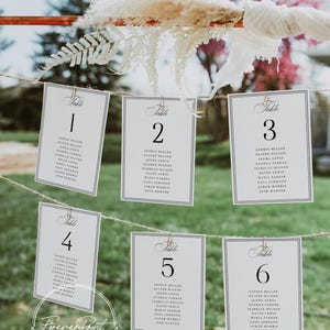 May include: Wedding table number cards hanging on a string. Each card is white with a black border and features a table number and guest names. The cards are held by small gold clips, with decorative floral elements.