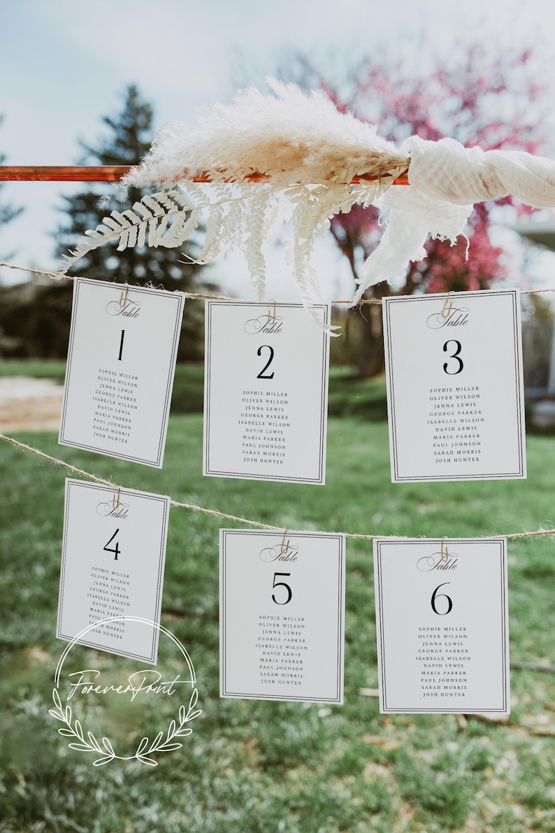 May include: Wedding table number cards hanging on a string. Each card is white with a black border and features a table number and guest names. The cards are held by small gold clips, with decorative floral elements.