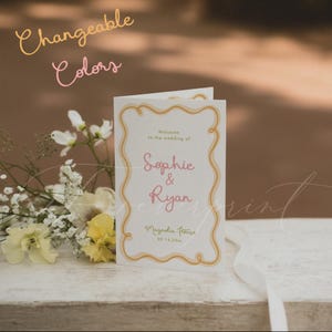 May include: A folded white wedding programme card featuring a wavy gold border, pink text reading Sophie & Ryan, and the text Welcome to the wedding of, Magnolia House, and 09.14.20xx. The text Changeable Colors appears in the upper left corner.