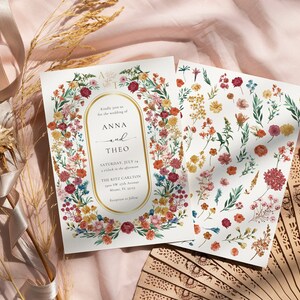 May include: Two wedding invitations with a floral design. The invitation features a gold oval frame with the text "Kindly join us for the wedding of Anna and Theo" and the date and location of the wedding. The invitation is surrounded by a wreath of colorful flowers. The second card features a variety of flowers and leaves.