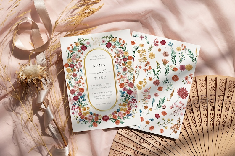May include: Two wedding invitations with a floral design. The invitation features a gold oval frame with the text "Kindly join us for the wedding of Anna and Theo" and the date and location of the wedding. The invitation is surrounded by a wreath of colorful flowers. The second card features a variety of flowers and leaves.