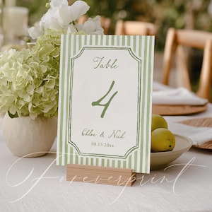 May include: A table number card with a green and white striped design. The card displays the word "Table" above the number "4", and the names "Chloe & Nick" with a date. The card is held in a wooden holder.