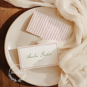 Op de afbeelding: Een gedekte tafel met twee naamkaartjes en een gestreepte kaart op een wit bord, gedrapeerd met een transparante stof. Het naamkaartje leest "Amelia Parker" in een scriptlettertype, met "Table 1" eronder.