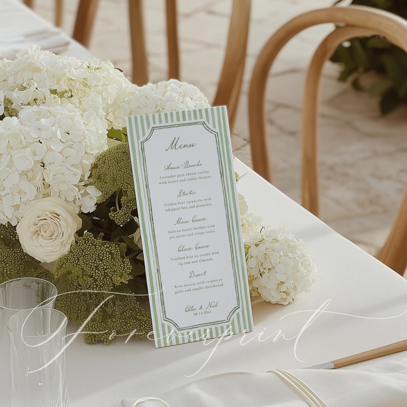 Op de afbeelding: Een menukaart met een groen-witte gestreepte rand, met een lijst van gerechten. Het menu staat op een witte tafel, omringd door witte bloemen en groen. Het menu bevat de namen van de gerechten en het bruidspaar.