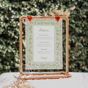 May include: A copper-colored metal frame holds a menu card with a floral design. The menu features a light green border with a pink gingham pattern and a list of courses: First Course, Main Course, and Dessert. The menu includes the text "Enjoy!"