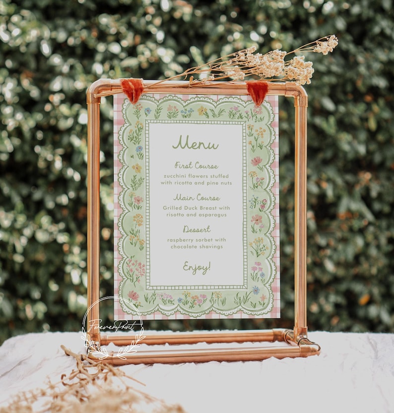 May include: A copper-colored metal frame holds a menu card with a floral design. The menu features a light green border with a pink gingham pattern and a list of courses: First Course, Main Course, and Dessert. The menu includes the text "Enjoy!"