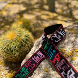 Peut inclure: Un bracelet noir tiss&eacute; avec du texte arabe brod&eacute; en couleur. Le bracelet est pos&eacute; sur un rocher avec un cactus en arri&egrave;re-plan.